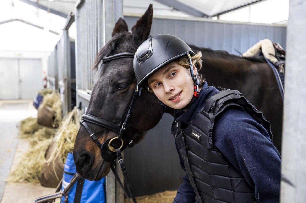 Ung ryttare med hjälm och skyddsväst bredvid en mörk häst i en stallgång