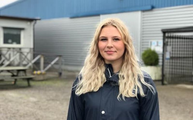 Ung kvinna med blont hår som ler mot kameran framför en byggnad